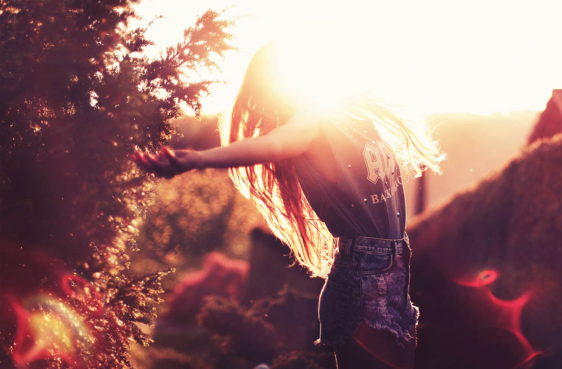 Woman standing outdoors during sunset with open arms, surrounded by nature. Her hair flows in the golden light, symbolizing freedom and connection with the environment.